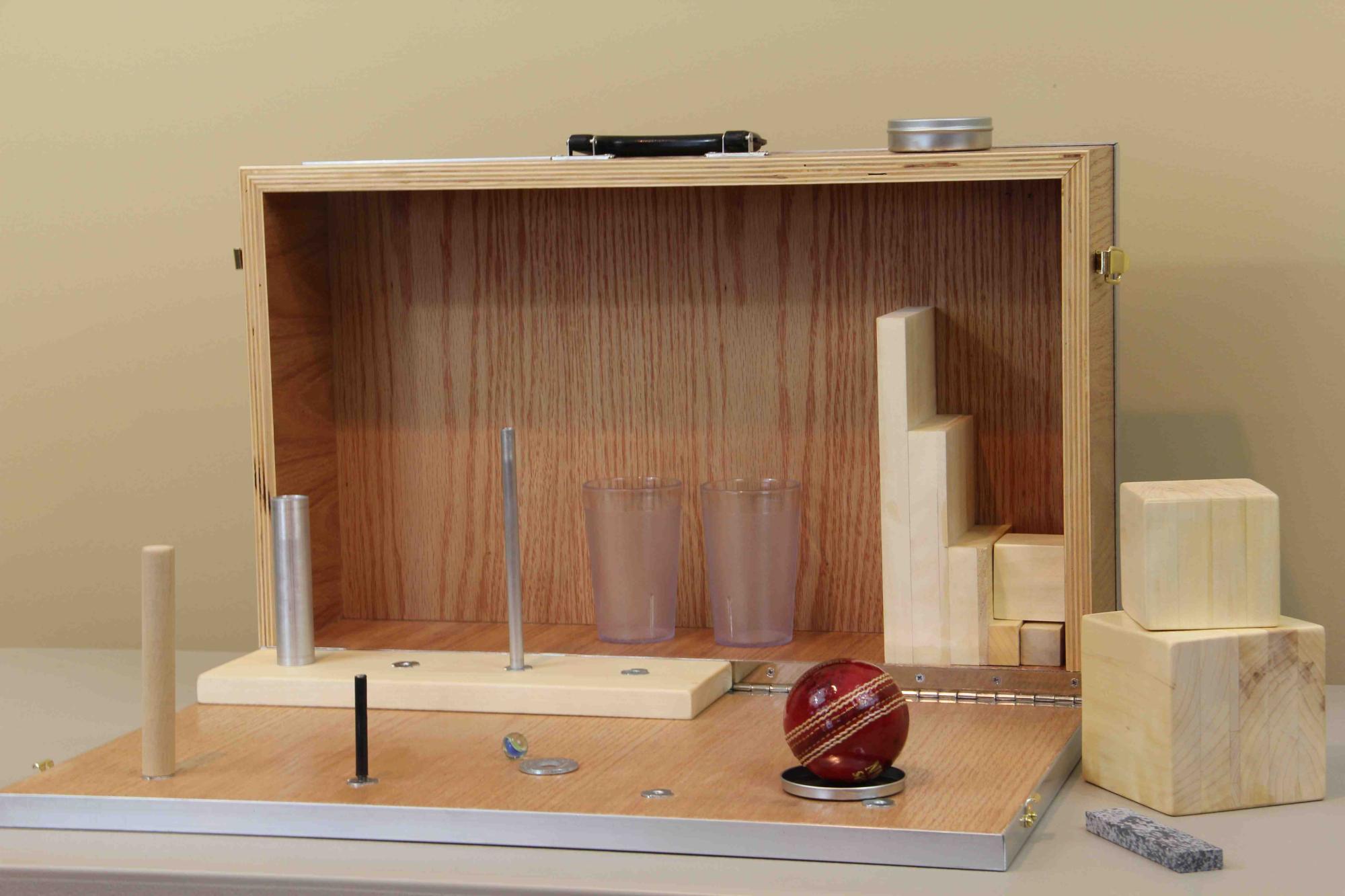 The Action Research Arm Test kit — wooden case with blocks, cylinders, marbles, and cricket ball for standardized upper-extremity assessment.